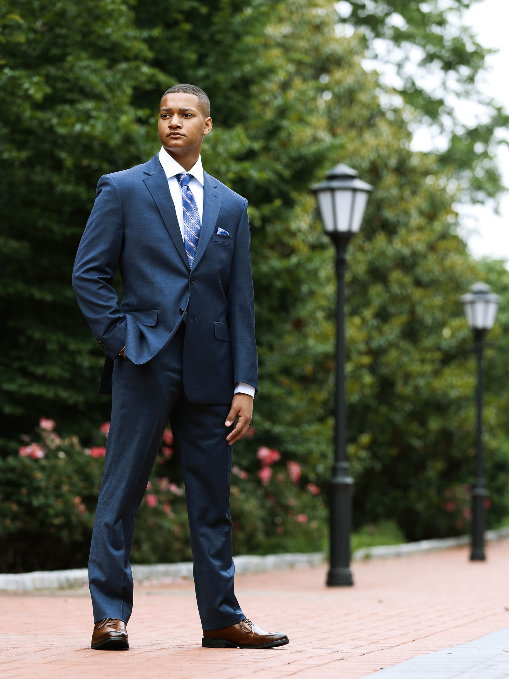 A young man dressed in a suit and tie stands on brick pavement with greenery and a light pole in the background -- Delaware senior portraits.